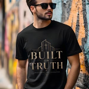 Puede incluir: Camiseta negra con la frase "BUILT ON TRUTH" en fuente beige, con un gráfico de un edificio y una cruz. El texto "JESUS CHRIST - OUR FOUNDATION" está debajo del texto principal.