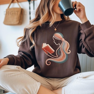 May include: A brown sweatshirt featuring a retro illustration of a person reading a book. The design includes a silhouette of a person with long, flowing hair in shades of blue, orange, and pink. The text "DIGITAL PR" is visible in the upper left corner.