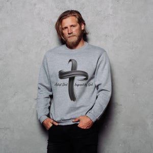 Puede incluir: Una sudadera gris con un gran diseño de cruz negra con pinceladas. El texto "Artist Dad Inspired by God" está debajo de la cruz. La sudadera es de manga larga y tiene cuello redondo.
