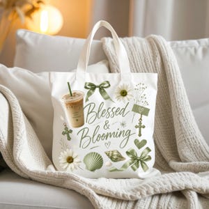 Puede incluir: Una bolsa de tela blanca con un estampado gráfico que incluye el texto Blessed & Blooming en letra cursiva verde. El diseño presenta un vaso de plástico con café helado, cintas verdes, margaritas blancas, conchas marinas verdes y cruces pequeñas.