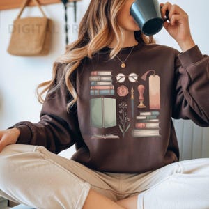 Puede incluir: Una sudadera marrón con un diseño gráfico de libros, gafas, un marcapáginas y una pluma. El diseño incluye pilas de libros de varios colores, un par de gafas redondas y una ilustración botánica. La sudadera tiene cuello redondo.