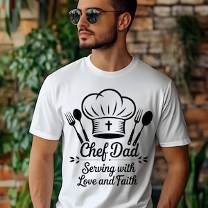 Puede incluir: Camiseta blanca con un diseño negro que incluye un gorro de chef, una cruz, un tenedor y una cuchara. El texto dice "Chef Dad Serving with Love and Faith". Cuello redondo.