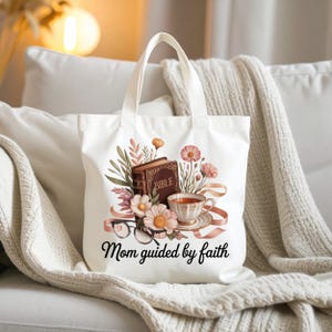 Puede incluir: Bolso tote blanco con un diseño floral que incluye una Biblia, una taza de té y gafas. El texto "Mom guided by faith" está impreso debajo de la ilustración. El bolso tiene dos asas blancas.