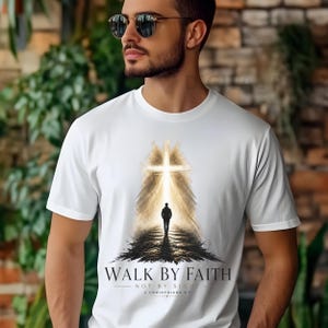Puede incluir: Camiseta blanca con un gráfico de una persona caminando hacia una cruz brillante. El texto "WALK BY FAITH" y "NOT BY SIGHT" está impreso debajo. La camiseta está hecha de un material suave.