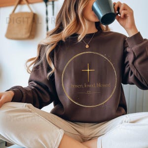 May include: A brown sweatshirt with a gold cross design inside a circle. The text "Chosen, loved, blessed" is below the cross. The sweatshirt is a solid color and has long sleeves.