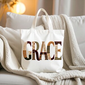 Puede incluir: Una bolsa de tela blanca con la palabra GRACE impresa en letras grandes y decorativas. Cada letra está rellena con un patrón único, incluyendo motivos florales, una pequeña cruz, un corazón y un símbolo de pez, en tonos tierra.
