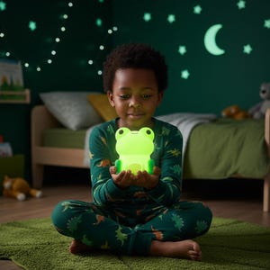 May include: A child holds a luminous green frog night light. The child is wearing green dinosaur-patterned pyjamas and sitting on a green rug. The background includes a bed, wall decorations, and a soft glow.