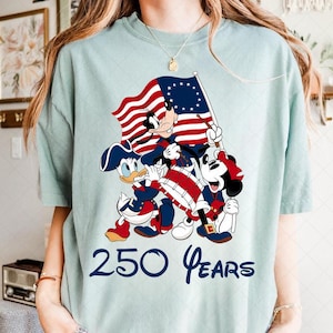 May include: Light blue t-shirt featuring Mickey Mouse, Donald Duck, and Goofy in patriotic attire, holding a flag. The flag has stars and stripes. The text "250 Years" is printed below the characters.