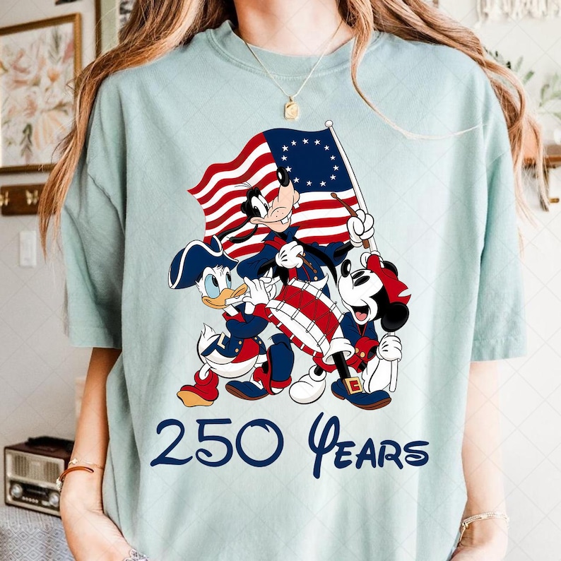 May include: Light blue t-shirt featuring Mickey Mouse, Donald Duck, and Goofy in patriotic attire, holding a flag. The flag has stars and stripes. The text "250 Years" is printed below the characters.