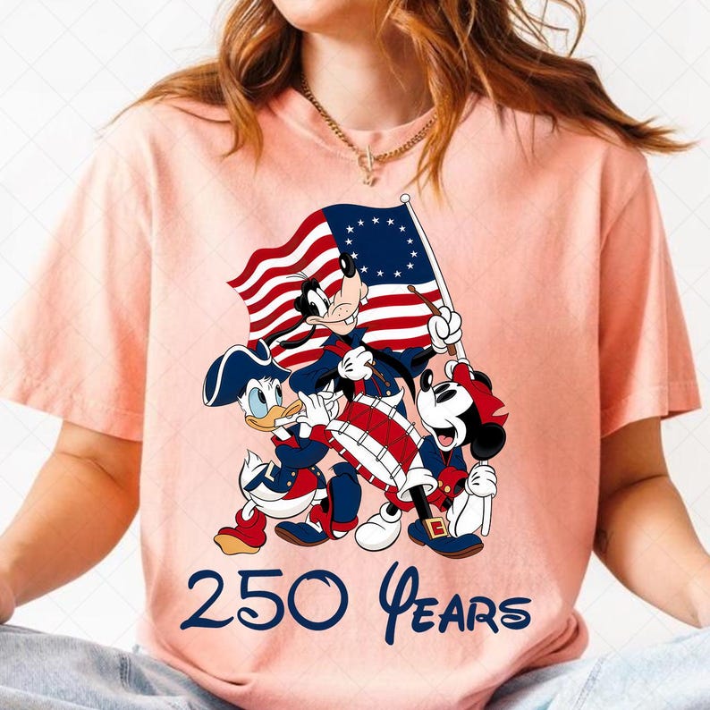 May include: A light pink t-shirt featuring a patriotic design with Mickey Mouse, Donald Duck, and Goofy in colonial attire, holding a flag and drum. The text "250 Years" is printed below the characters.