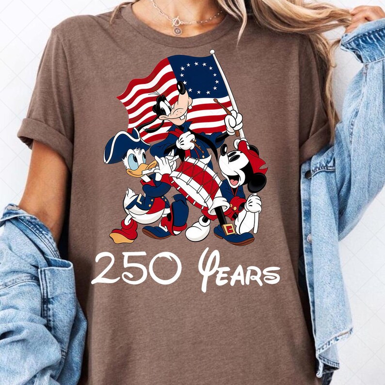 May include: Brown t-shirt featuring Mickey Mouse, Donald Duck, and another mouse character in patriotic attire, holding a flag and drum. The flag has red and white stripes with a blue field of stars. The text "250 Years" is printed below the characters.