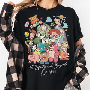 May include: Black t-shirt featuring a colorful graphic of Toy Story characters, including Woody, Buzz Lightyear, and Jessie. The words "Toy Story Land" are at the top, with "To Infinity and Beyond! Est. 1995" at the bottom.