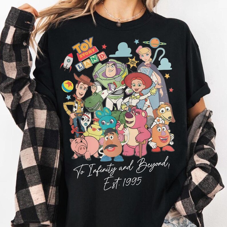 May include: Black t-shirt featuring a colorful graphic of Toy Story characters, including Woody, Buzz Lightyear, and Jessie. The words "Toy Story Land" are at the top, with "To Infinity and Beyond! Est. 1995" at the bottom.