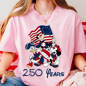 May include: Pink t-shirt featuring a patriotic design with cartoon characters, including Donald Duck, Goofy, and Mickey Mouse, holding a flag and playing a drum. The text "250 Years" is printed in blue at the bottom.