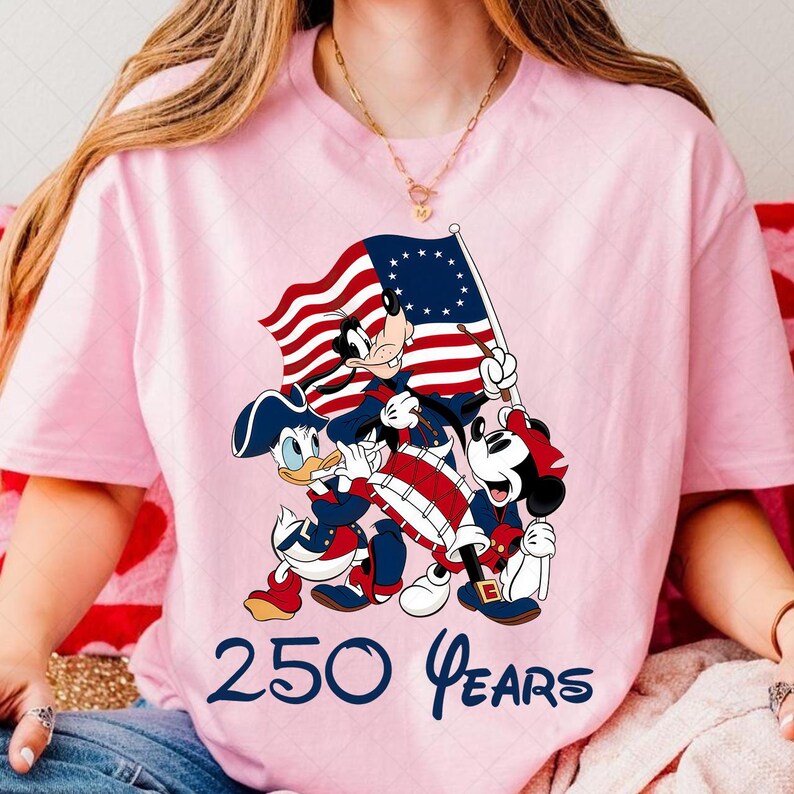 May include: Pink t-shirt featuring a patriotic design with cartoon characters, including Donald Duck, Goofy, and Mickey Mouse, holding a flag and playing a drum. The text "250 Years" is printed in blue at the bottom.