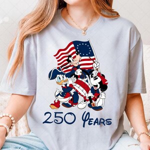 May include: Light blue t-shirt featuring a patriotic design with Mickey Mouse, Donald Duck, and Goofy dressed in Revolutionary War attire, holding a flag and drum. The text "250 Years" is printed below the characters.