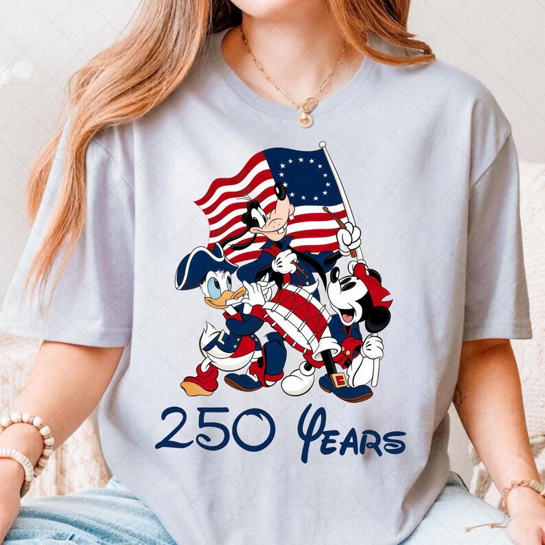 May include: Light blue t-shirt featuring a patriotic design with Mickey Mouse, Donald Duck, and Goofy dressed in Revolutionary War attire, holding a flag and drum. The text "250 Years" is printed below the characters.