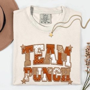 Puede incluir: Camiseta color crema con la frase "TEAM PUNCH" en un diseño de patchwork. Las letras son en tonos marrón, blanco y naranja, con detalles de estrellas. También se ve un collar dorado con un colgante de estrella.