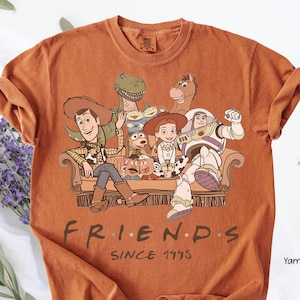 May include: Orange t-shirt featuring a graphic of Toy Story characters on a couch. The design includes Woody, Buzz Lightyear, and other characters. The text "FRIENDS" and "SINCE 1995" are printed below the graphic.