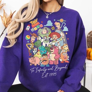 May include: A purple sweatshirt featuring a colorful graphic of characters from the animated film "Toy Story." The design includes the text "Toy Story Land" and "To Infinity and Beyond, Est. 1995."