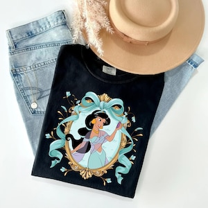 Puede incluir: Camiseta negra con una ilustración de princesa de dibujos animados en un marco dorado y una cinta azul. La camiseta se combina con vaqueros azul claro y un sombrero color canela. La camiseta tiene el texto "COMFORT COLORS".