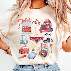 May include: Cream-colored t-shirt featuring a Cars-themed design. The design includes various cartoon cars, a traffic light, checkered flags, and the word "Cars" in a red and silver emblem. Additional elements include stars and a trophy.