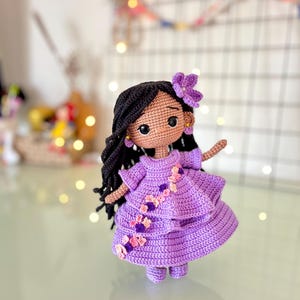 May include: A handmade crochet doll with dark hair and a purple dress. The doll has a flower in its hair and is adorned with small floral accents on the dress. The doll is standing on a reflective surface.