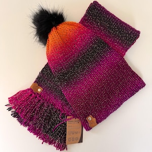 May include: A hand-knitted hat and scarf set. The hat has a black pom-pom and a gradient of orange, red, and black. The scarf is a matching gradient of pink, purple, and black with fringe. Both items have leather tags.