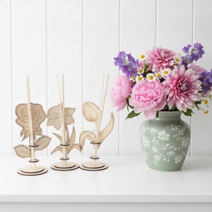 Könnte beinhalten: Drei helle, blumenförmige Vermehrungsstationen aus Holz mit durchsichtigen Glasröhrchen, die jeweils einen Holzstab enthalten, neben einer hellgrünen Keramikvase mit weißem Blumenmuster und einem Strauß aus rosa Dahlien.