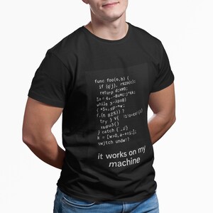 May include: Black t-shirt with white text and code-like graphics. The text reads "it works on my machine" below a block of code. The shirt is a classic crew neck style.