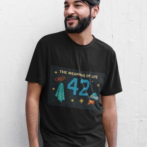 Könnte beinhalten: Schwarzes T-Shirt mit einem grafischen Design mit dem Text "THE MEANING OF LIFE" und der Zahl "42" in Blau. Das Design umfasst ein UFO, ein Handtuch und Sterne. Das T-Shirt wird von einer Person getragen.