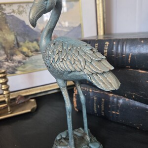 Pode incluir: Uma estatueta de flamingo em pé com um acabamento de pátina azul-esverdeada desgastada. A ave tem um pescoço longo e curvo, penas texturizadas detalhadas no corpo e apoia-se numa base pequena e irregular que imita o solo natural.