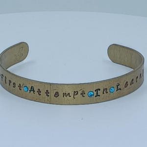 May include: A brass-colored cuff bracelet with the inscription "first attempt in learning" and small turquoise-colored accents. The bracelet has a slightly curved shape and is open at the ends.