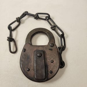 May include: An antique, oval-shaped padlock with a dark, aged finish. The lock features a central rectangular plate and several rivets. A matching dark chain is attached to the lock, creating a secure closure. The lock is likely made of metal.