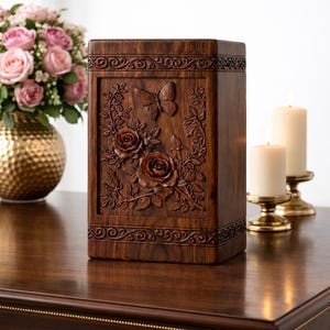 Op de afbeelding: Een donkerbruine houten urn met gesneden bloemen- en vlinderdetails. De rechthoekige urn heeft rozen en een vlinder in het midden, met decoratieve randen. De urn staat op een donkere houten tafel, met kaarsen en bloemen op de achtergrond.