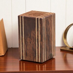Peut inclure: Une boîte en bois brun foncé avec des rayures verticales de couleur claire. La boîte est rectangulaire et repose sur une surface en bois foncé. Deux livres et une petite plante en pot sont visibles en arrière-plan.