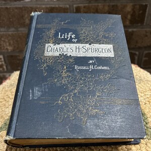 May include: A vintage book titled "Life of Charles H. Spurgeon" by Russell H. Conwell. The book has a dark blue cover with gold floral designs and text. The title is in a rectangular white box. The book is slightly worn, showing its age.