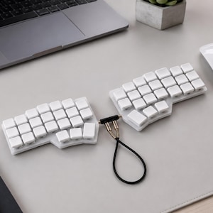 May include: A split ergonomic keyboard featuring two separate white sections with blank white keycaps. The two halves are connected by a short black cable with metallic connectors, creating a distinct layout designed for two-handed typing.