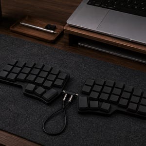 May include: A black split ergonomic mechanical keyboard with blank keycaps, connected by a short black cable with silver connectors. The keyboard consists of two separate halves, each featuring a grid layout of square keys, positioned on a dark gray mat.