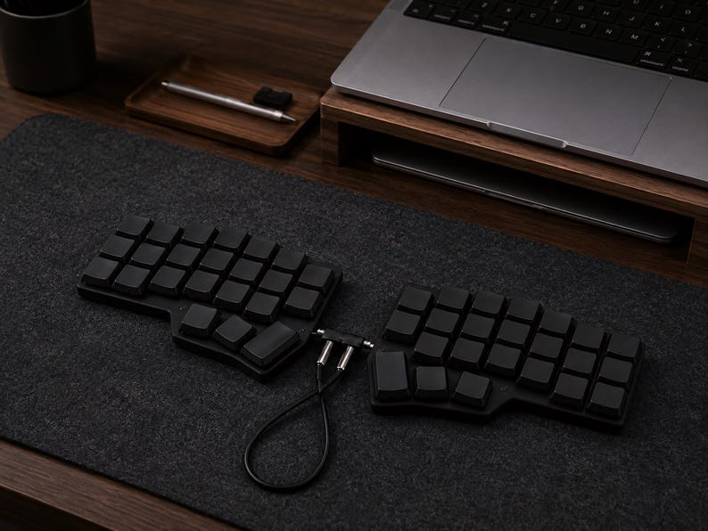 May include: A black split ergonomic mechanical keyboard with blank keycaps, connected by a short black cable with silver connectors. The keyboard consists of two separate halves, each featuring a grid layout of square keys, positioned on a dark gray mat.