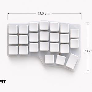 May include: A white ergonomic split keyboard with blank keycaps, measuring 13.5 cm in width and 9.3 cm in height. The device features a non-standard, contoured layout with a transparent base and visible screws. The logo ART is in the corner.