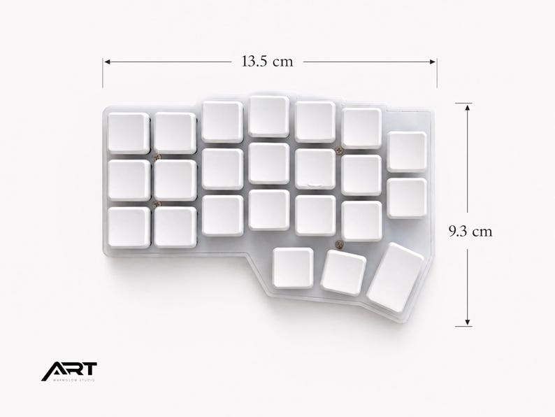 May include: A white ergonomic split keyboard with blank keycaps, measuring 13.5 cm in width and 9.3 cm in height. The device features a non-standard, contoured layout with a transparent base and visible screws. The logo ART is in the corner.