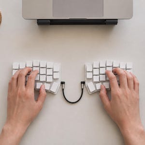 May include: A split ergonomic mechanical keyboard featuring two separate white keycap sections connected by a black cable. The keyboard layout is ortholinear with blank white keys, positioned on a flat surface in front of a laptop computer.