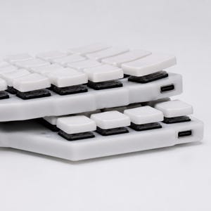 May include: Two stacked white split mechanical keyboard halves featuring white keycaps and black switches. Each unit has a visible USB-C port on the side. The design is low-profile with a contoured layout, showcasing the internal switch mechanism.