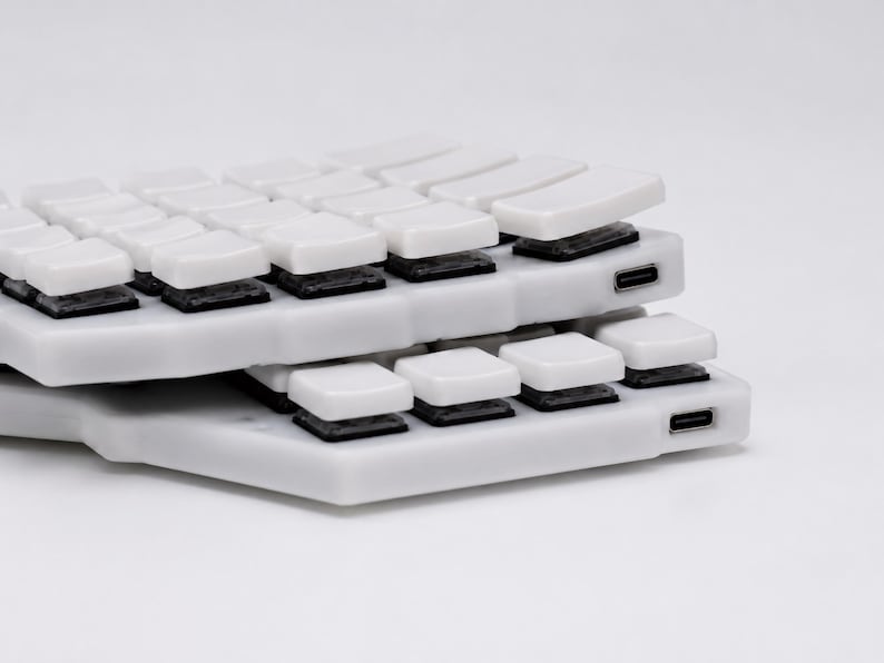 May include: Two stacked white split mechanical keyboard halves featuring white keycaps and black switches. Each unit has a visible USB-C port on the side. The design is low-profile with a contoured layout, showcasing the internal switch mechanism.