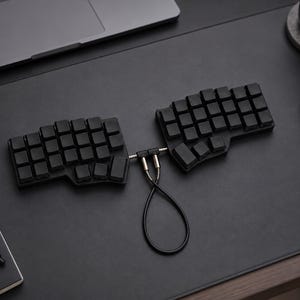 May include: A black split ergonomic mechanical keyboard consisting of two separate halves connected by a short, looped black cable with metallic silver connectors. The keyboard features a blank, uniform keycap layout without any visible lettering.