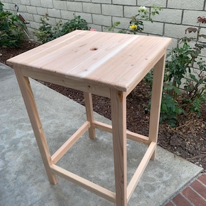 May include: A tall, square-topped wooden table constructed from light-coloured, unfinished wood planks. The table features four straight legs connected by lower support rails on all sides and a small, circular hole centred in the tabletop surface.