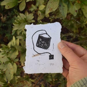 Puede incluir: Una taza de cerámica blanca con un asa negra y el texto "But First, Coffee" en negro, con una fuente de estilo manuscrito. La taza está sobre una superficie blanca.