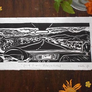 May include: Black and white linocut print depicting a car interior with a road leading into a landscape. The print includes the text "Good Times Remembered". The artwork is on textured paper, surrounded by flowers.