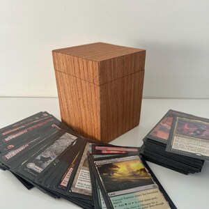 May include: A wooden card storage box with a lid, made of light brown wood. Several decks of trading cards are fanned out in front of the box, with some cards stacked to the right. The cards have various colorful illustrations.